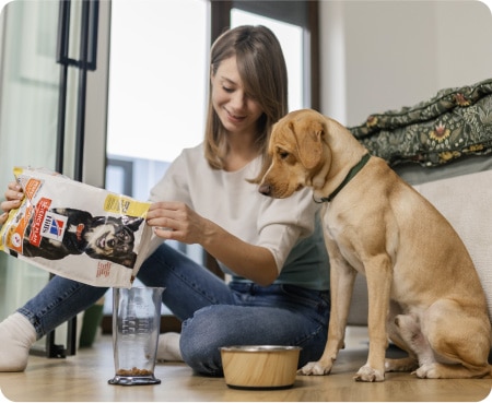 Nainen kaataa Hill’s Pet Nutrition -ruokaa mitta-astiaan, kun koira seuraa tilannetta tarkkaavaisesti.